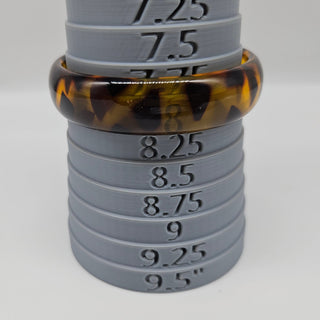A tortoiseshell bangle displayed on a gray plastic sizer stand with size markings, photographed against a light gray background.