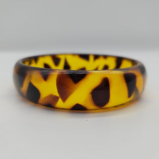 A glossy tortoiseshell-patterned bangle with a vibrant yellow base and dark brown and black mottled spots, shown on a neutral light gray background.