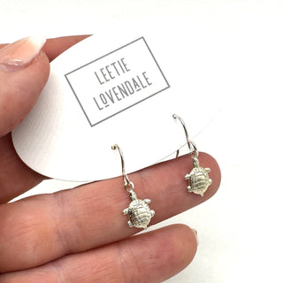 Silver turtle earrings held between fingers with 'Leetic Lovendale' branding in the background.