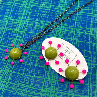 OlIve green earrings with hot pink beads radiating from center with posts and matching necklace pendant on a chain on a green and blue plaid background.