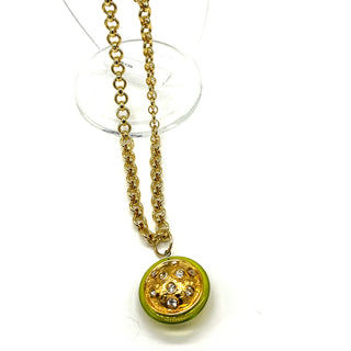 A gold chain necklace with a round pendant featuring a lime green outer ring and a gold-toned center decorated with small clear rhinestones, displayed on a clear stand against a white background.