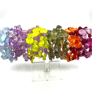 A set of colorful beaded bracelets displayed on a clear acrylic stand against a white background. The bracelets feature various translucent and opaque beads in shades of purple, yellow, green, orange, and red, with some spiral and swirl bead designs.