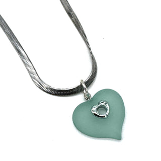 A silver snake chain necklace with a heart-shaped frosted lucite pendant in a muted teal green color, featuring a small silver heart charm with a metallic center, set against a plain white background.