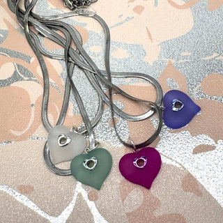 Four heart-shaped pendant necklaces with silver chains, displayed on a patterned beige and silver surface, each featuring a small silver heart cutout in the center.