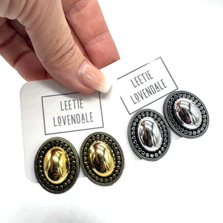 A close-up of a hand holding white cards displaying a pair of oval stud earrings. The earrings have shiny gold and silver oval centers surrounded by dark antiqued metal beaded borders.