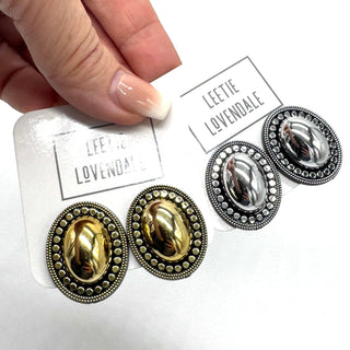 A close-up of a hand holding a white card displaying two pairs of oval stud earrings on a white background. The pairs are gold-toned and silver-toned with ornate beaded borders and smooth oval centers.