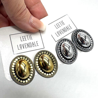 A close-up of a hand holding a white card printed with the brand name 'LEETIE LOVENDALE' above a pair of oval stud earrings. The card also shows a second pair of matching earrings.
