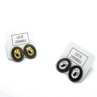 Two pairs of oval stud earrings on white display cards. One pair features gold metal with a textured border, and the other pair features silver metal with a textured border.