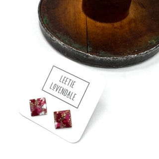 A pair of square stud earrings with a deep pink and burgundy resin design and gold foil flakes, displayed on a white card labeled 'LEETIE LOVENDALE' next to a rustic wooden stand.