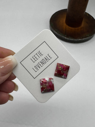 A hand holds a white display card featuring two small square stud earrings with a pink, red, and white marbled pattern and gold foil flecks. The card is branded 'LEETIE LOVENDALE'.