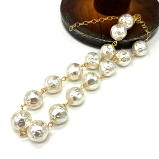 A strand necklace made of irregular, hammered white pearls connected by delicate gold chains, displayed on a clean white background with a dark wooden stand partially visible in the upper right.