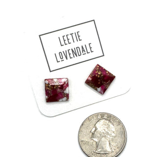 A pair of square glass stud earrings with a deep magenta-pink and white marbled pattern and gold flecks, displayed on a white card labeled 'LEETIE LOVENDALE' next to a US quarter for scale.