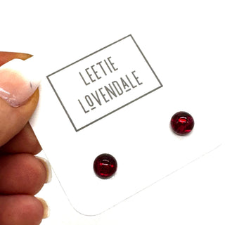 A close-up of a hand holding a white display card with the brand name 'LEETIE LOVENDALE' printed on it. The card holds two small, round, dark red stud earrings.