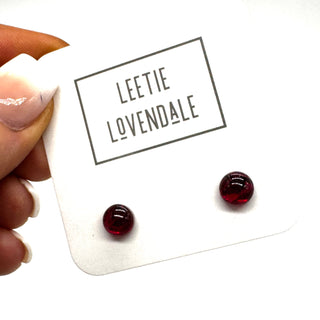 A hand holding a white display card with the brand name 'LEETIE LOVENDALE' and two small round dark red stud earrings attached, photographed against a white background.