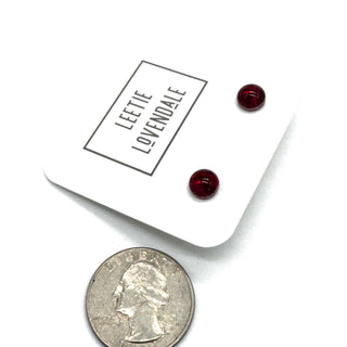 Two small round dark red stud earrings displayed on a white branded card next to a U.S.
