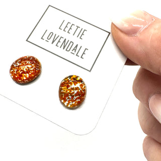 A close-up of a hand holding a white display card with two oval stud earrings. The card is branded 'LEETIE LOVENDALE' and the studs are clrar glass with orange flecks on a white background.