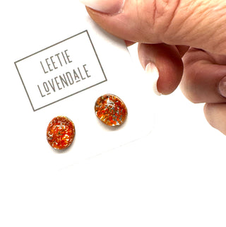 A close-up of a hand holding a white card with the brand name 'LEETIE LOVENDALE' above two oval orange stud earrings, on a white background.