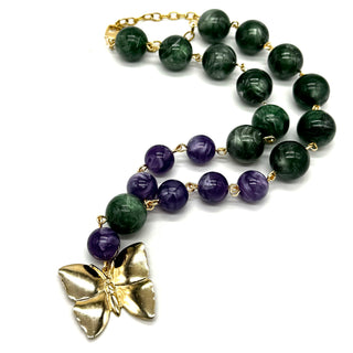 A beaded necklace featuring dark green lucite beads, purple amethyst beads, and a textured gold butterfly pendant, displayed on a clean white background.
