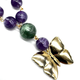 A close-up of a beaded necklace featuring purple and green marbled beads, gold-toned links, and a large textured gold butterfly pendant on a white background.