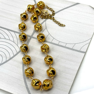 A gold-tone beaded necklace with irregular, textured spherical beads connected by delicate chains, displayed on a white textured card with gray line art.