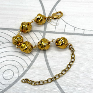A gold-toned metal bracelet featuring textured, rounded beads and a matching chain extender, displayed on a white background with a gray circular graphic pattern.