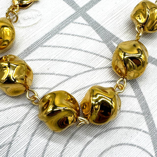 A close-up of a gold-tone beaded bracelet with irregular, organic-shaped beads connected by small links, laid on a white textured surface with gray curved line patterns.
